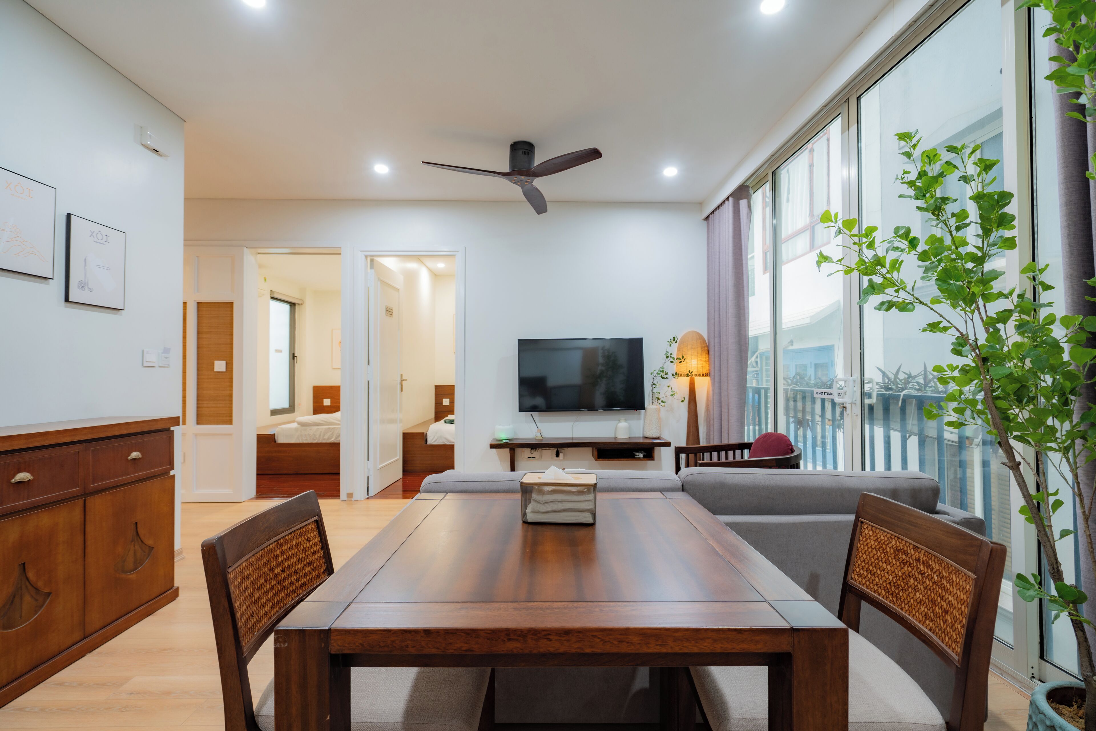 A modern living room with a wooden dining table, a sofa, a TV, and a balcony with a plant.