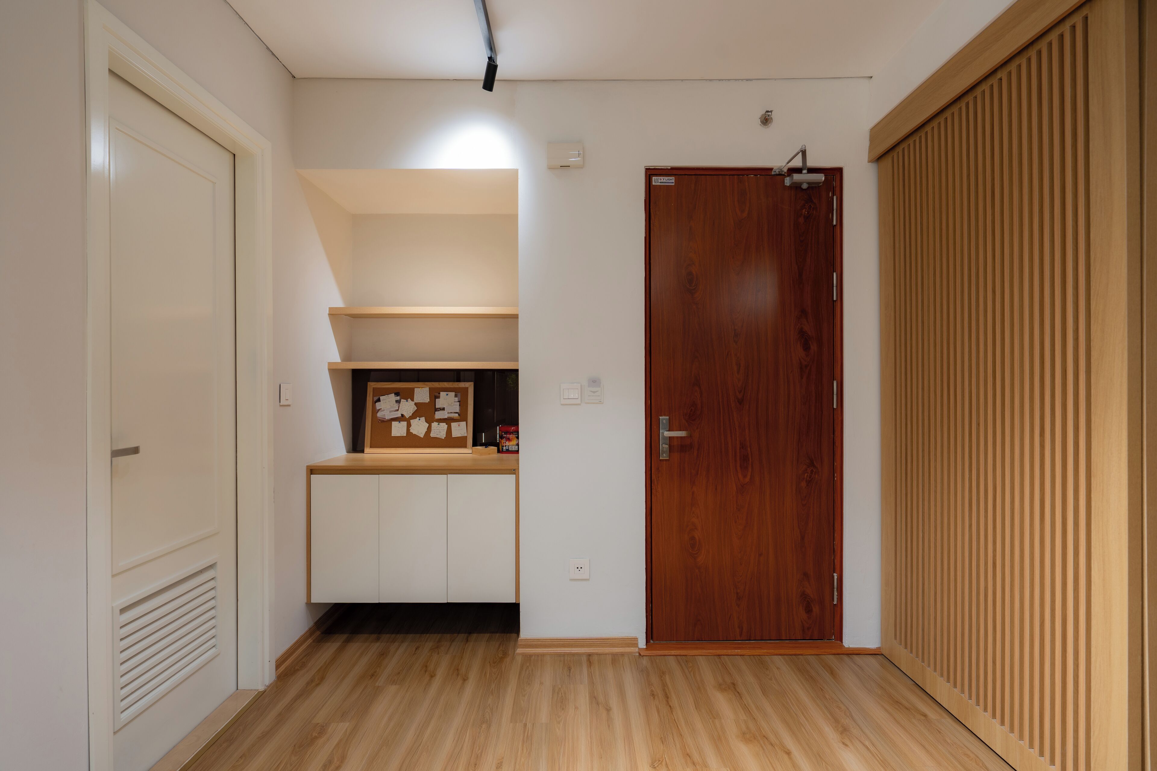 Un pasillo con piso de madera, una puerta blanca, un armario de madera y una pequeña zona de cocina con armarios y una encimera.