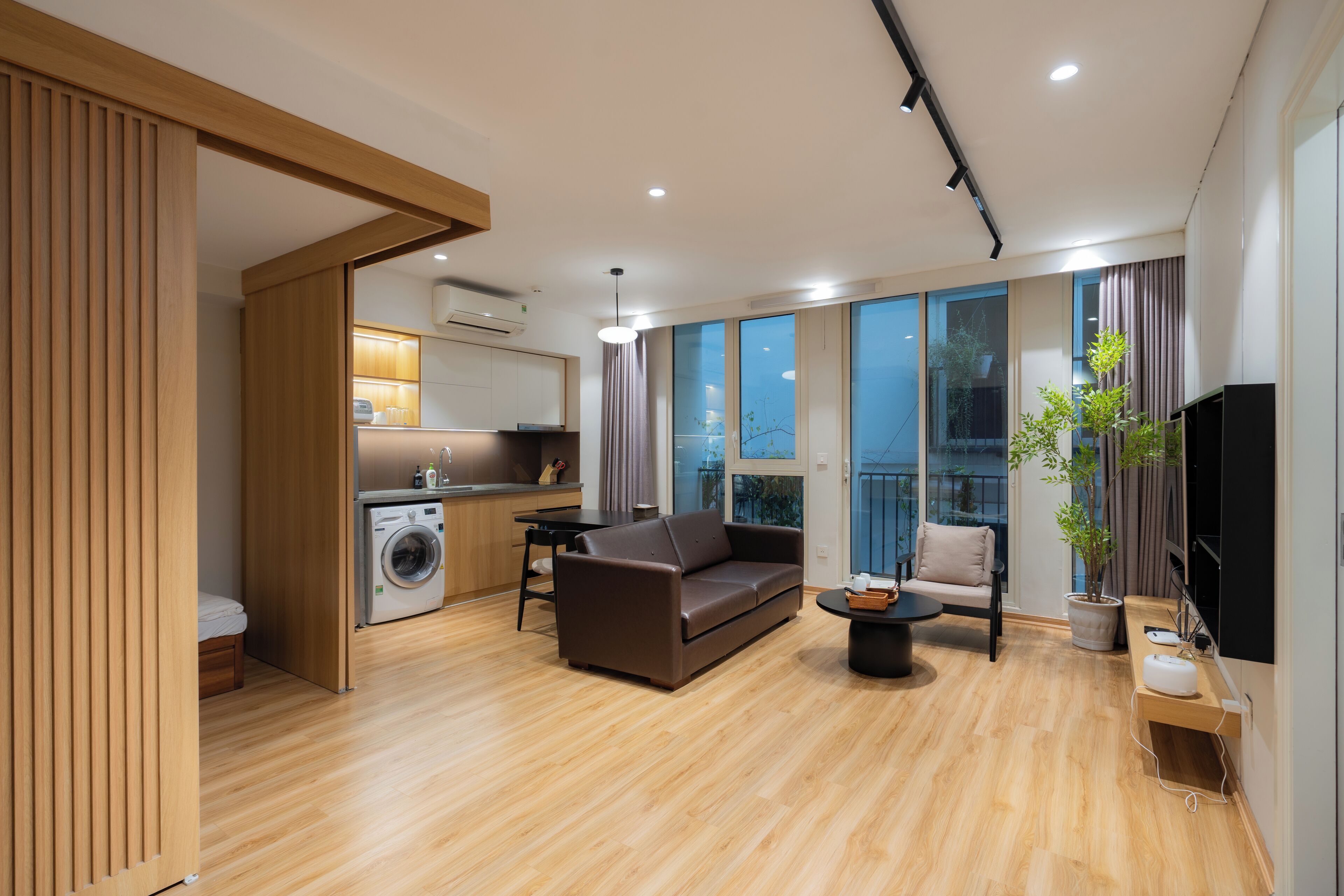 A modern living room with a sofa, a coffee table, and a TV. There is a kitchen area with a washing machine and a dining table.