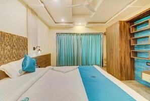 Une chambre d’hôtel avec un lit, un ventilateur de plafond, une table de chevet, une armoire en bois et une fenêtre avec des rideaux.