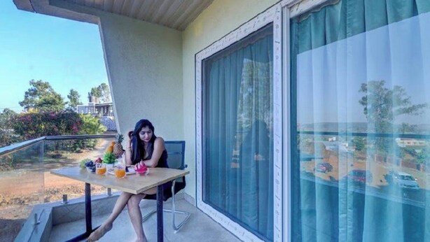 Un balcon avec une table, une chaise et une personne assise. On aperçoit une fenêtre avec des rideaux, ainsi qu’une vue sur un parking et des arbres en arrière-plan.