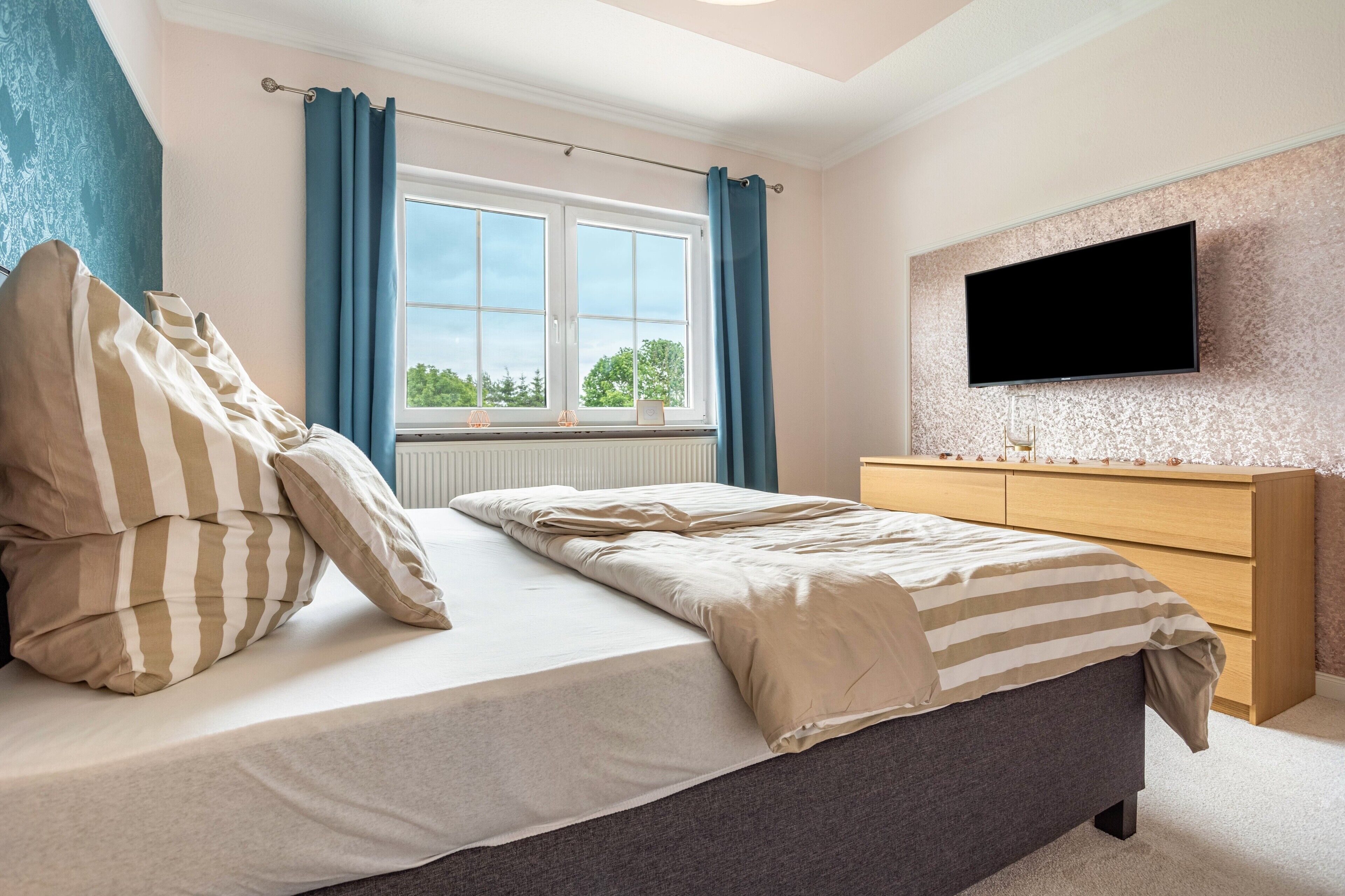 Ein Schlafzimmer mit einem Bett, einer Kommode, einem an der Wand montierten Fernseher und einem Fenster mit Vorhängen.