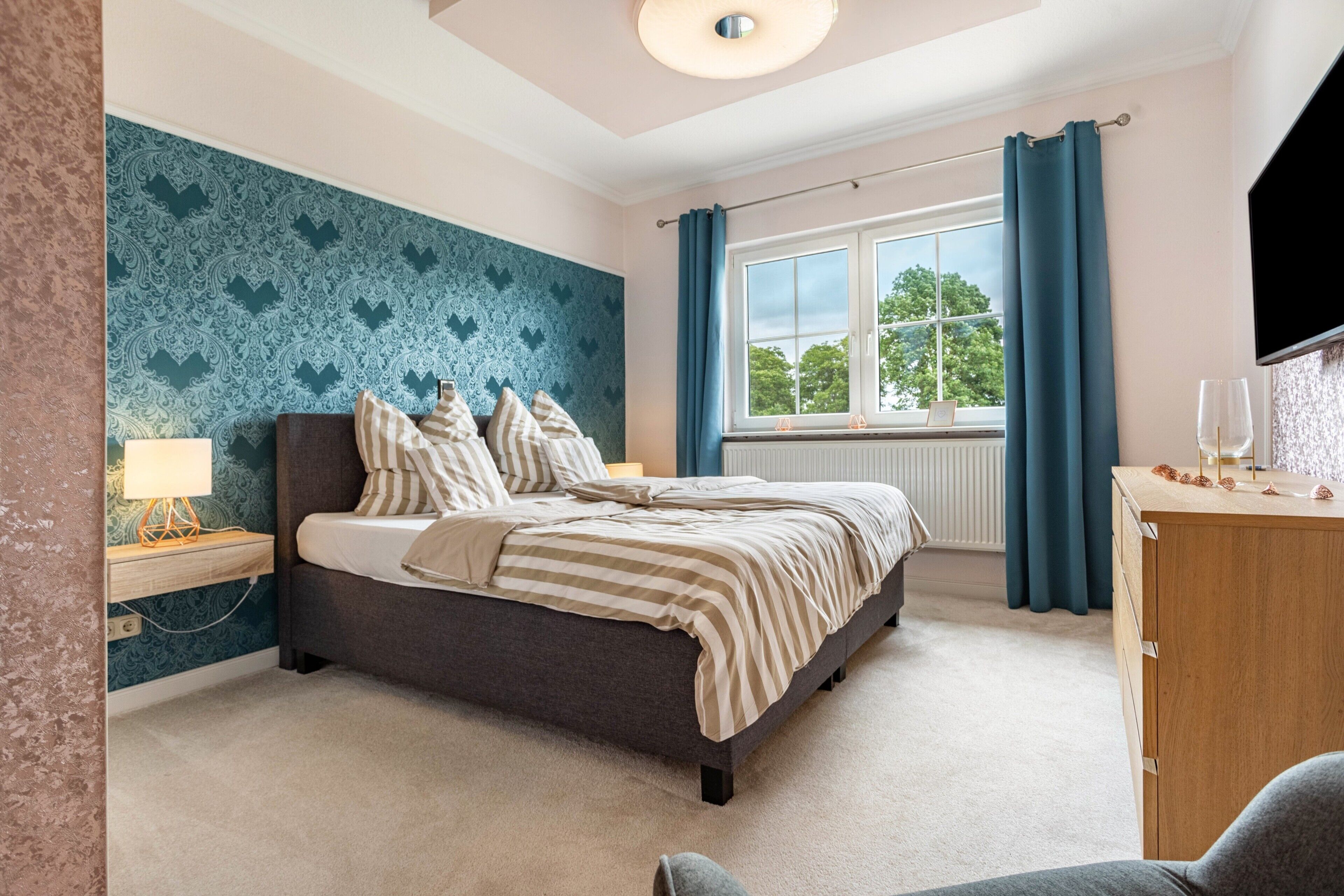Une chambre à coucher comprenant un lit, une table de chevet, une commode, une fenêtre avec des rideaux et un mur recouvert d’un papier peint à motifs.