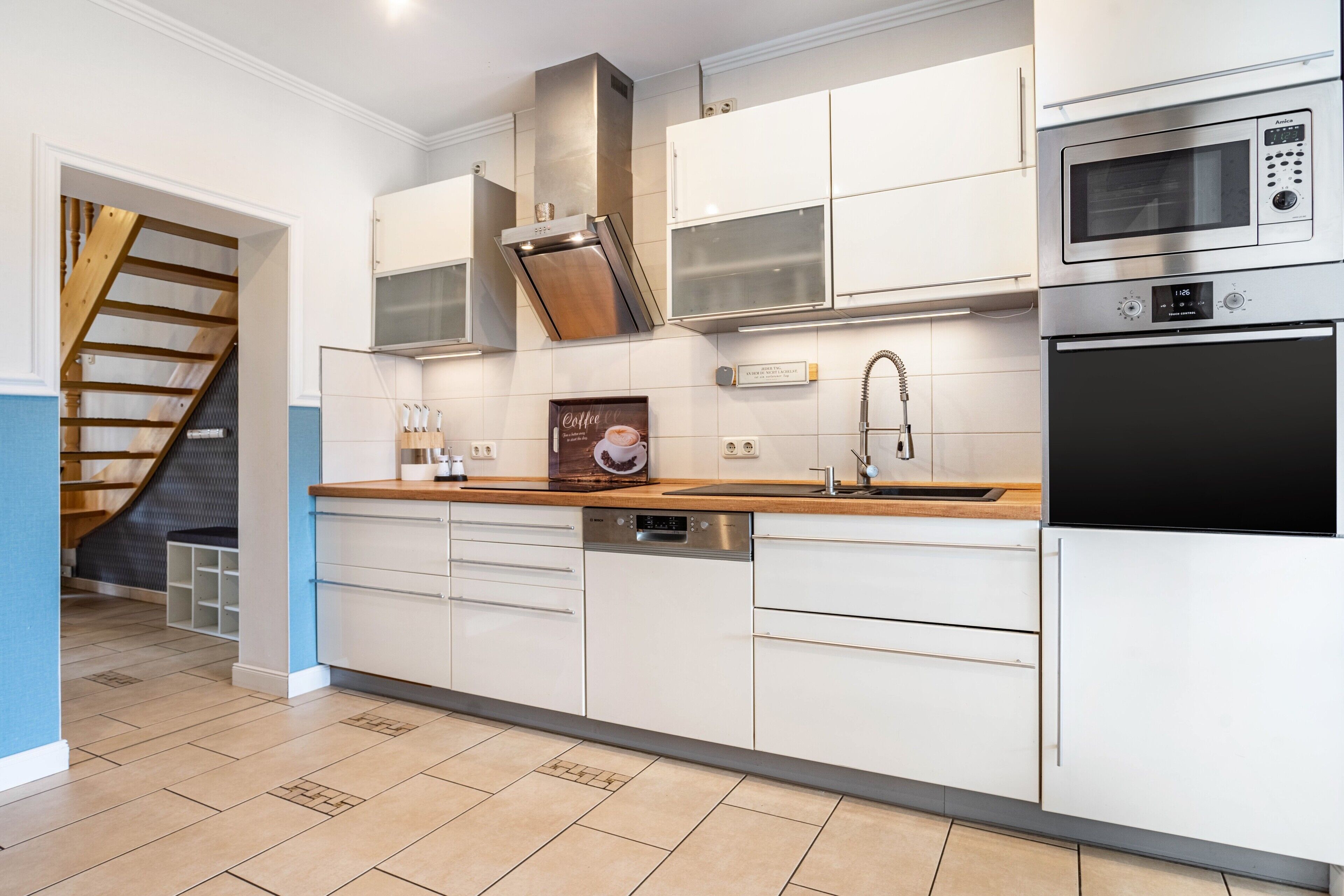 A modern kitchen with white cabinetry, stainless steel appliances, a dishwasher, and a wooden staircase.
