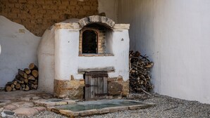 A rustic stone oven with a wooden door, surrounded by a gravel area and stacked firewood.