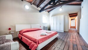 A bedroom with a wooden floor, a bed with a red bedspread, a white bedside table with a lamp, and a wardrobe.