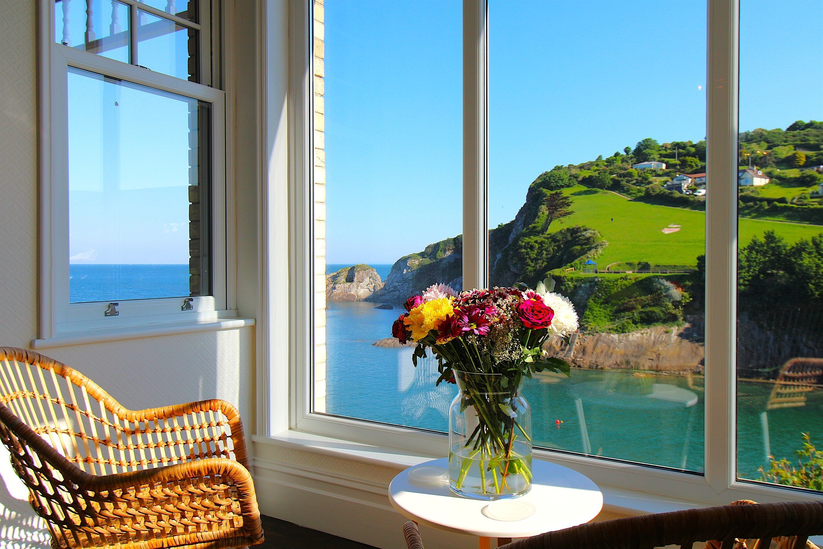 Ein Korbstuhl neben einem Fenster mit Blick auf eine Küstenlandschaft und ein Blumenstrauß auf einem kleinen Tisch.