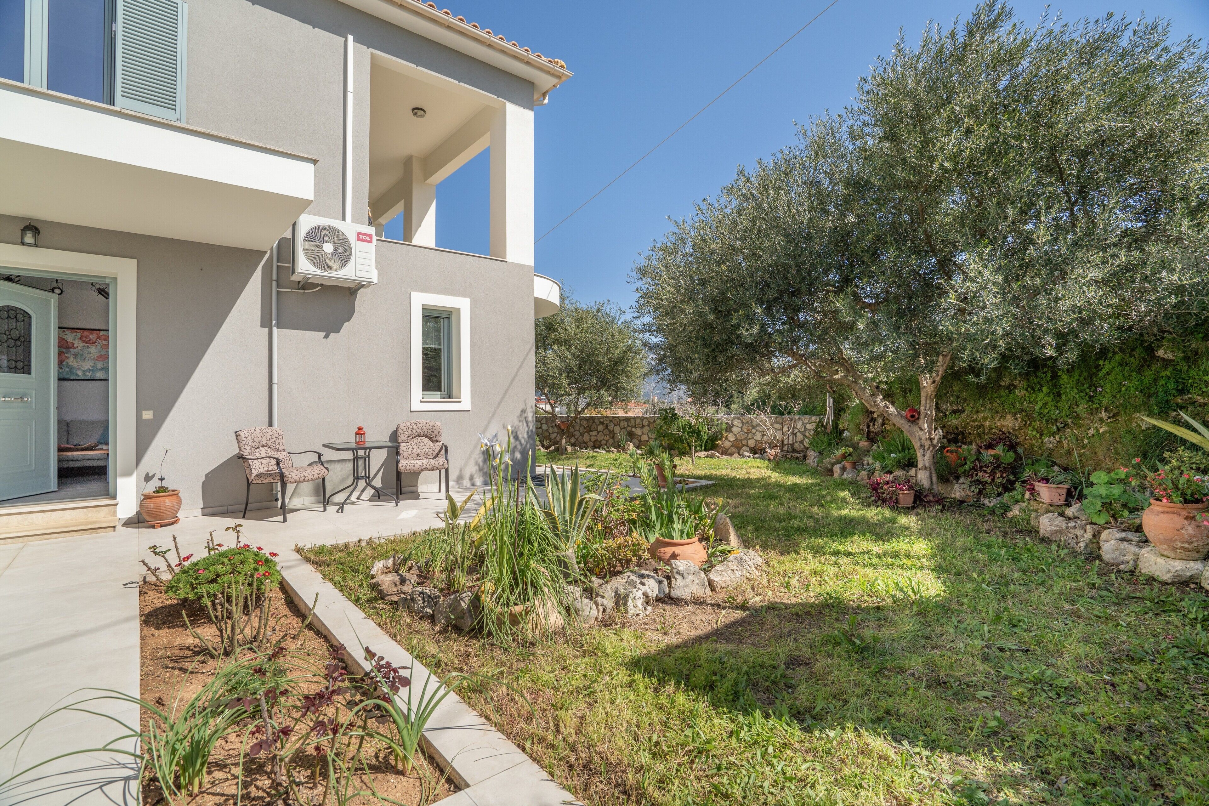 A modern two-story house with a balcony, outdoor seating, and a garden with various plants and a tree.