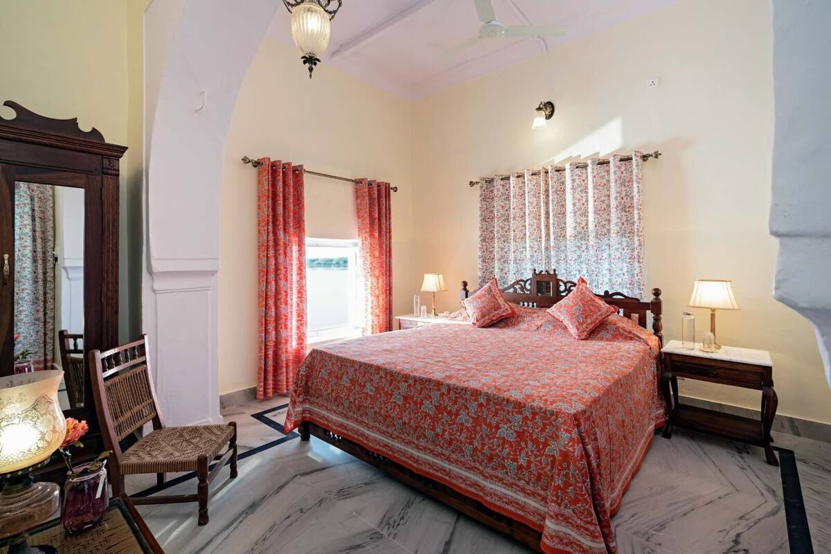 Une chambre à coucher avec un grand lit, deux tables de chevet avec des lampes, une chaise en bois, un miroir et une fenêtre avec des rideaux.