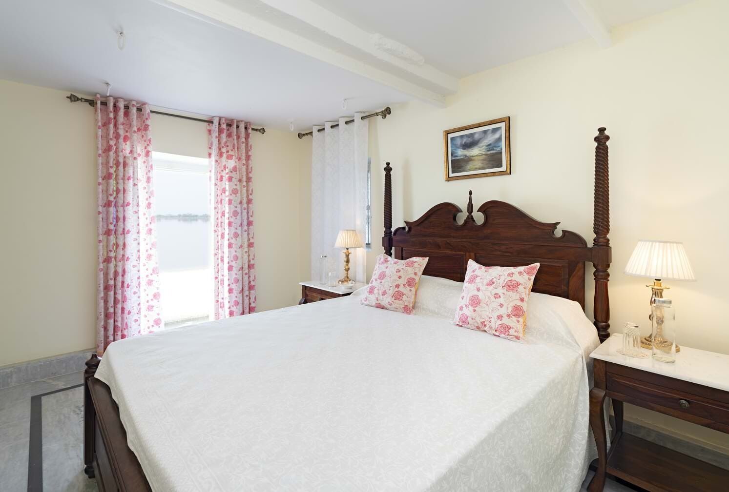 Una cama bien hecha con almohadones florales, cabecera de madera y dos mesitas de noche con lámparas.