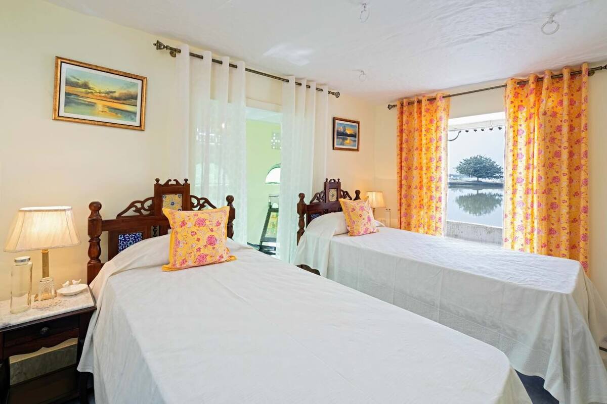 Une chambre d’hôtel avec deux lits, une table de chevet avec une lampe, un tableau au mur et une fenêtre avec des rideaux.