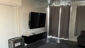 A modern living room with a flat-screen TV mounted on a white wall, a small heater, and a dark sofa. - Cabin and horse paddock for rent (Bective)