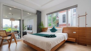 Ein modernes Hotelzimmer mit einem großen Bett, einer Sitzecke mit Sessel und Tisch, einer Schiebetür zum Außenbereich und einem großen Fenster mit Blick auf Gebäude und Grünflächen.
