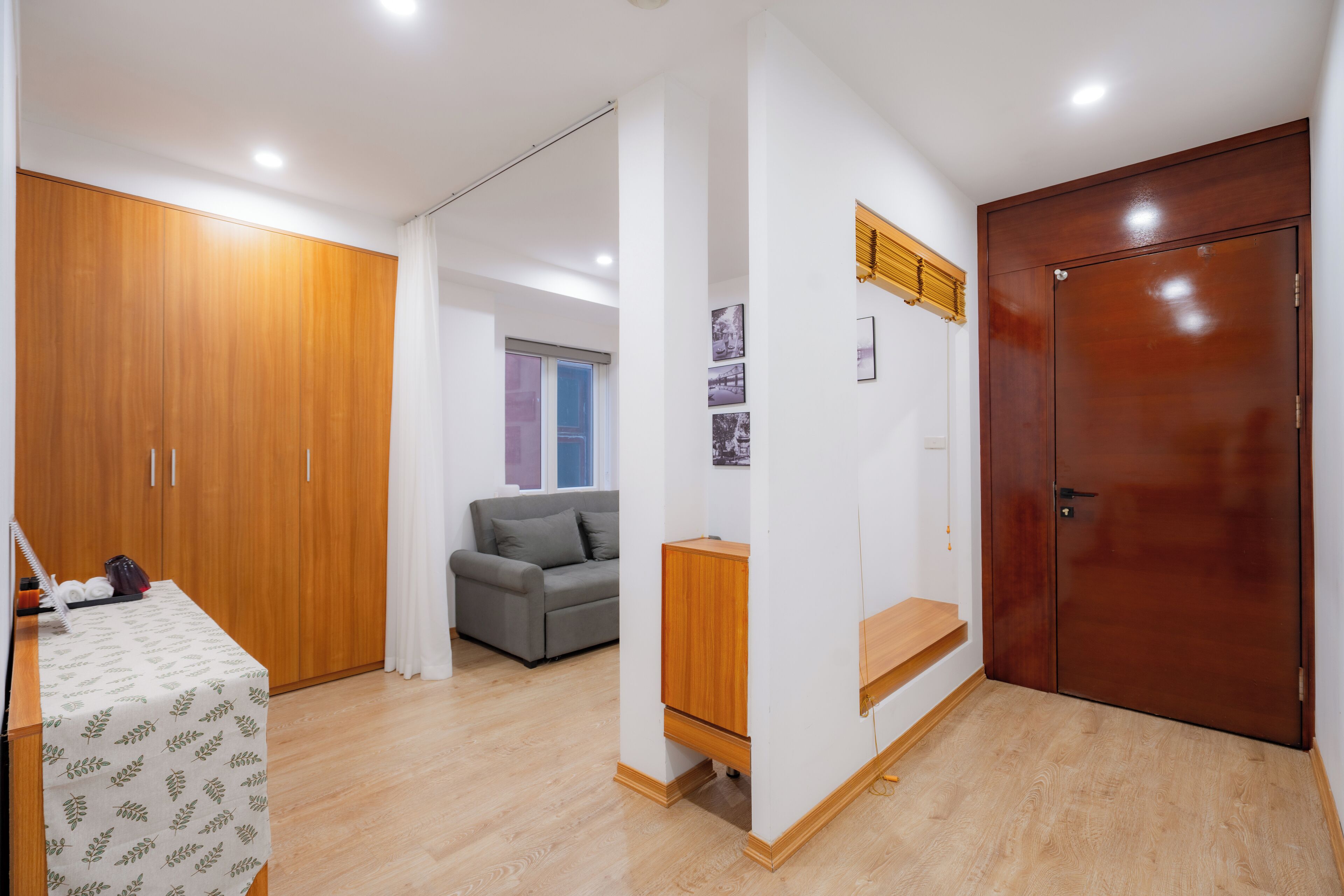 A room with a wooden wardrobe, a grey sofa, a small table with a tablecloth, and a wooden cabinet.