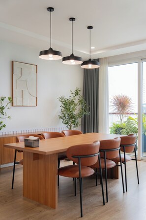 Un espace de salle à manger moderne, avec une table et des chaises en bois, des luminaires suspendus et une vue sur l’extérieur.
