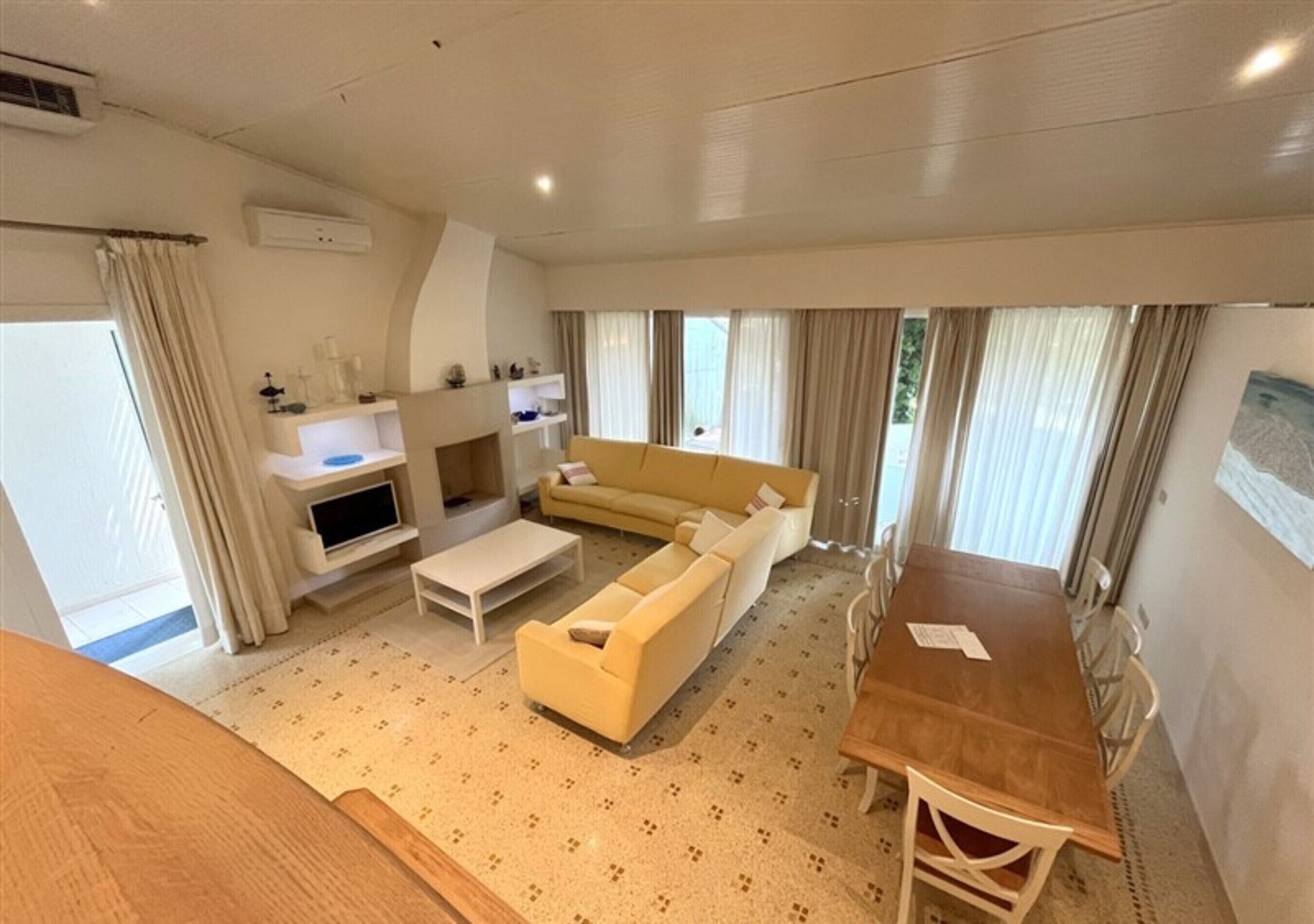 A modern living room with a sofa, coffee table, and a dining table with chairs. There is a fireplace, a TV, and a window with curtains.