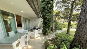 An outdoor dining area with a glass table and white chairs, surrounded by greenery and a tree. - Charming and Tastefully Furnished Villa Steps From the sea (Lignano Sabbiadoro)