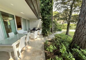 An outdoor dining area with a glass table and white chairs, surrounded by greenery and a tree. - Charming and Tastefully Furnished Villa Steps From the sea (Lignano Sabbiadoro)