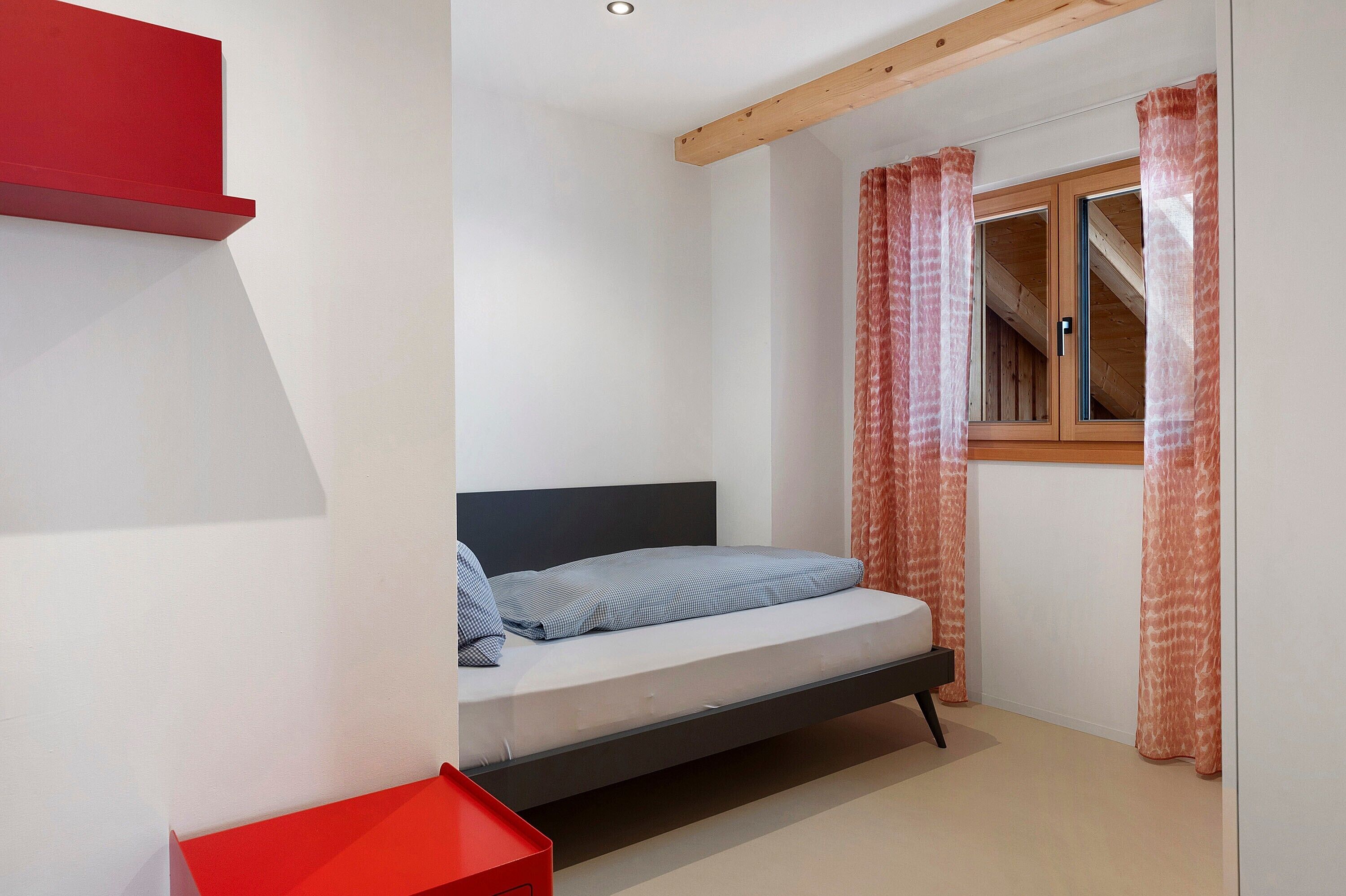 Ein Schlafzimmer mit einem Bett, einem roten Regal, einem Fenster mit Vorhängen und einer Holzbalkendecke.