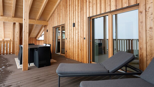 Eine Holzdiele mit Balkon, einem Tisch mit Stühlen und einem Liegestuhl.
