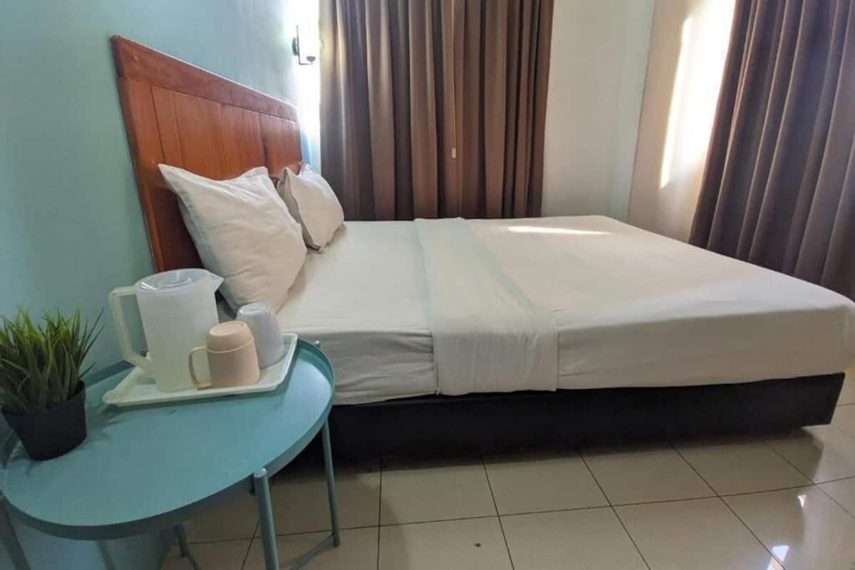 A neatly made bed with white linens, a wooden headboard, and a small table with a plant, a pitcher, and two cups.
