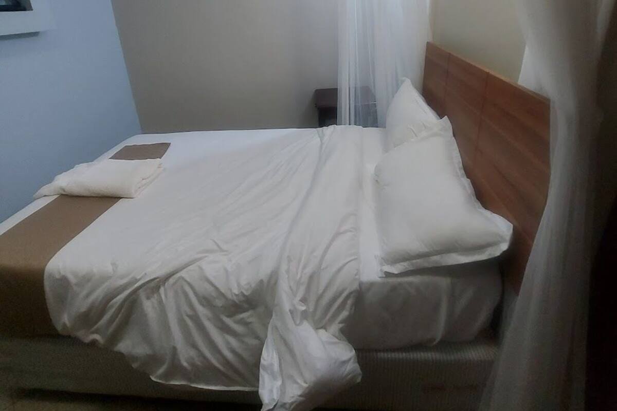 Un lit simple avec une tête de lit en bois, une literie blanche et un rideau blanc transparent.