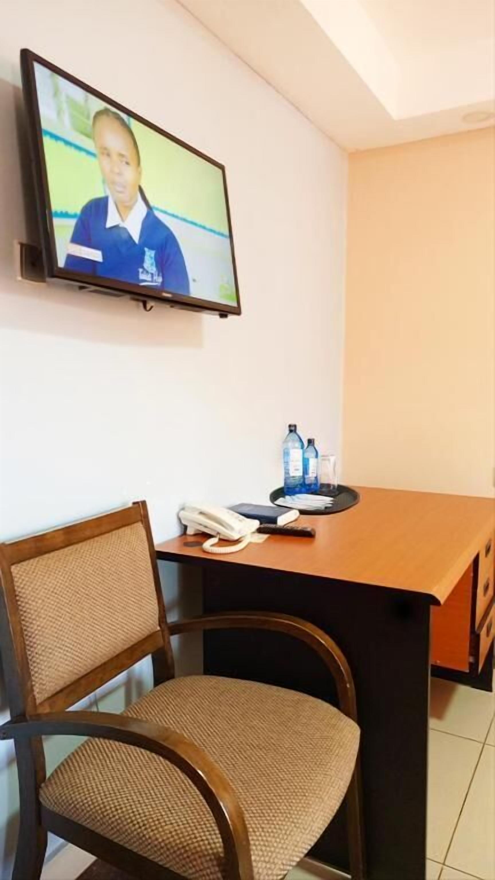 A hotel room with a television mounted on the wall, a wooden desk with a chair, a telephone, and two bottles of water.