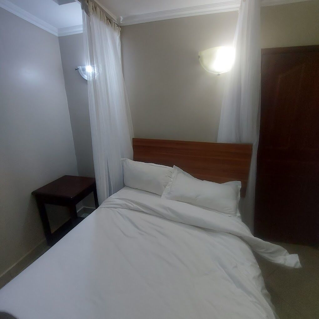 Una cama bien hecha con ropa de cama blanca, cabecera de madera, mesita de noche y lámpara fijada a la pared.