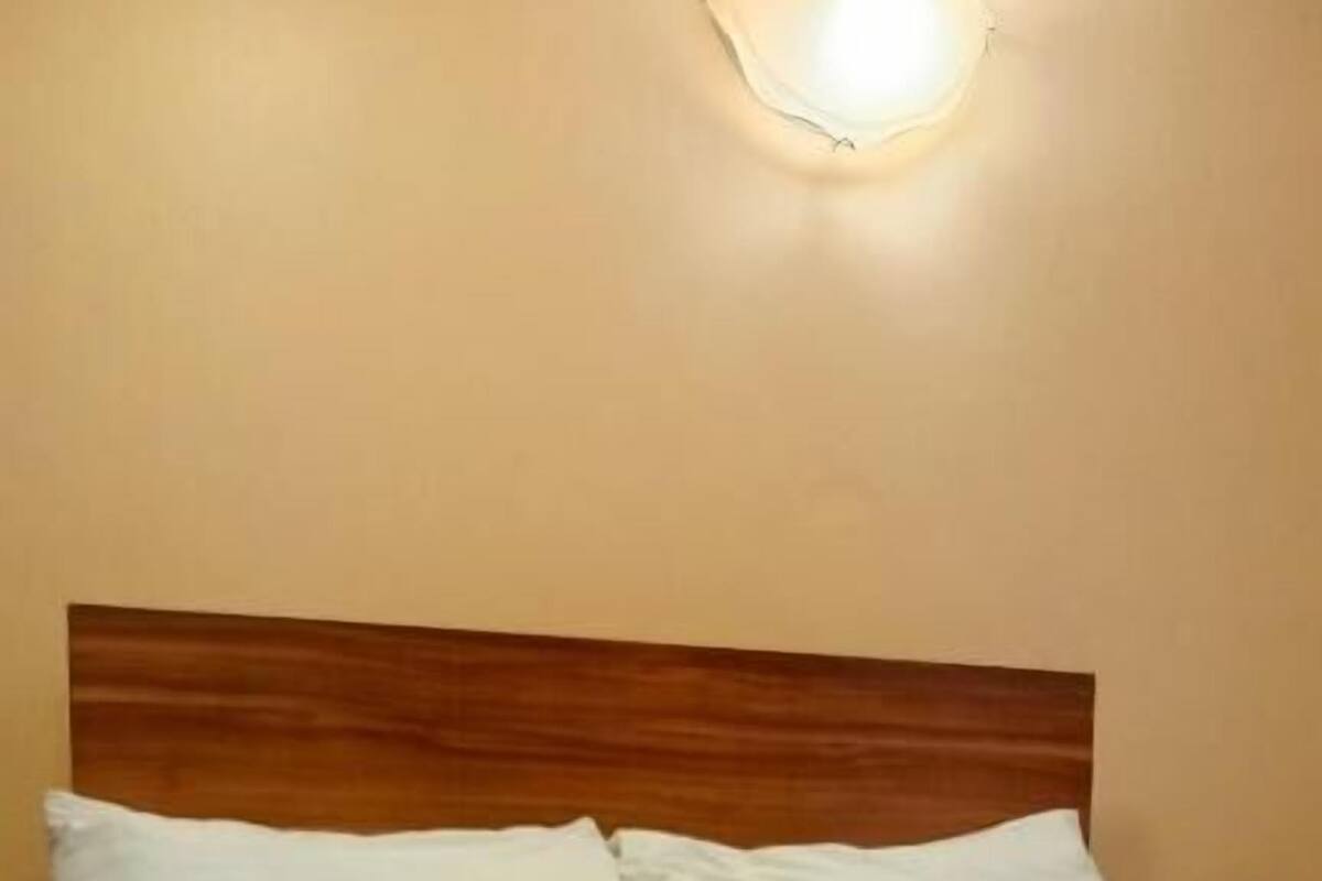 Un lit bien fait, avec du linge de lit blanc et un plaid marron, une tête de lit en bois et un luminaire fixé au mur.