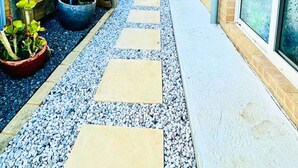 Ein schmaler Pfad mit trittfesten Steinen, begrenzt von einem blauen Zaun und einem Backsteinhaus.