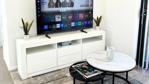 Ein modernes Wohnzimmer mit einem Flachbildfernseher auf einer weißen Unterhaltungsanlage, einem runden Couchtisch mit Zeitschriften, einer Uhr und einem abstrakten Kunstwerk an der Wand.