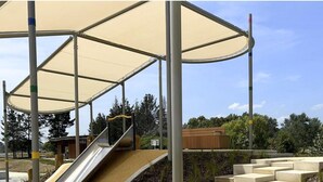 Ein Spielplatz mit Rutsche und Treppe, umgeben von schattigen Bereichen und Grünflächen.