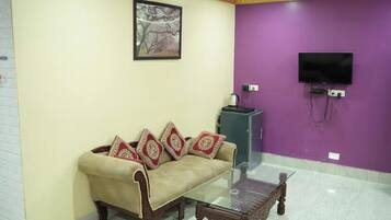 A living room with a sofa, a glass coffee table, a television mounted on a purple wall, and a ceiling fan.