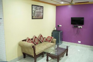 A living room with a sofa, a glass coffee table, a television mounted on a purple wall, and a ceiling fan.