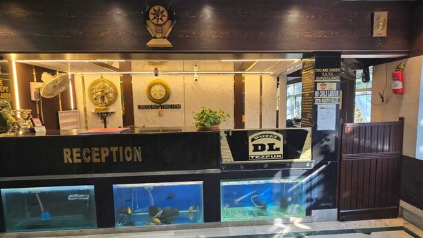 A reception desk with a sign reading "RECEPTION" and "DL TEZPUR." There are clocks on the wall and a sign indicating "CHECK OUT TIME 1PM." - Hotel DL (Tezpur)