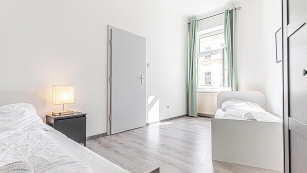 A bedroom with two beds, a bedside table with a lamp, a wardrobe, and a window with curtains. - Cozy 2BR Apartment Near Prater Danube (Vienna)