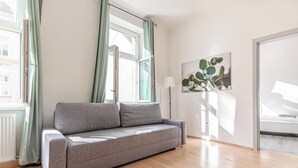 A modern living room with a grey sofa, a large window with curtains, a floor lamp, and a bed in the background. - Cozy 2BR Apartment Near Prater Danube (Vienna)
