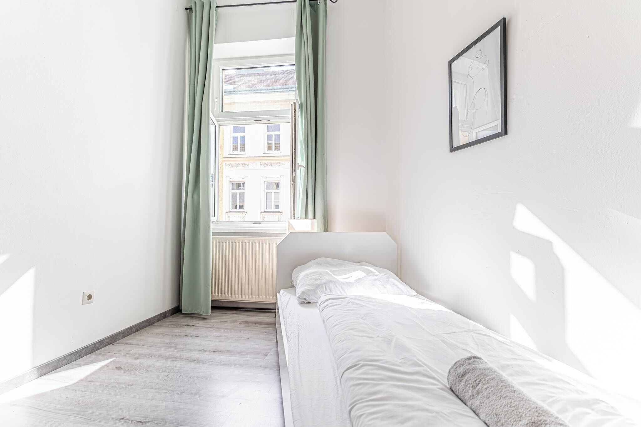 Ein kleiner, heller Raum mit einem Einzelbett, einem fenster mit Vorhängen und einem gerahmten Bild an der Wand.