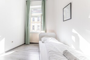 Ein kleiner, heller Raum mit einem Einzelbett, einem fenster mit Vorhängen und einem gerahmten Bild an der Wand.