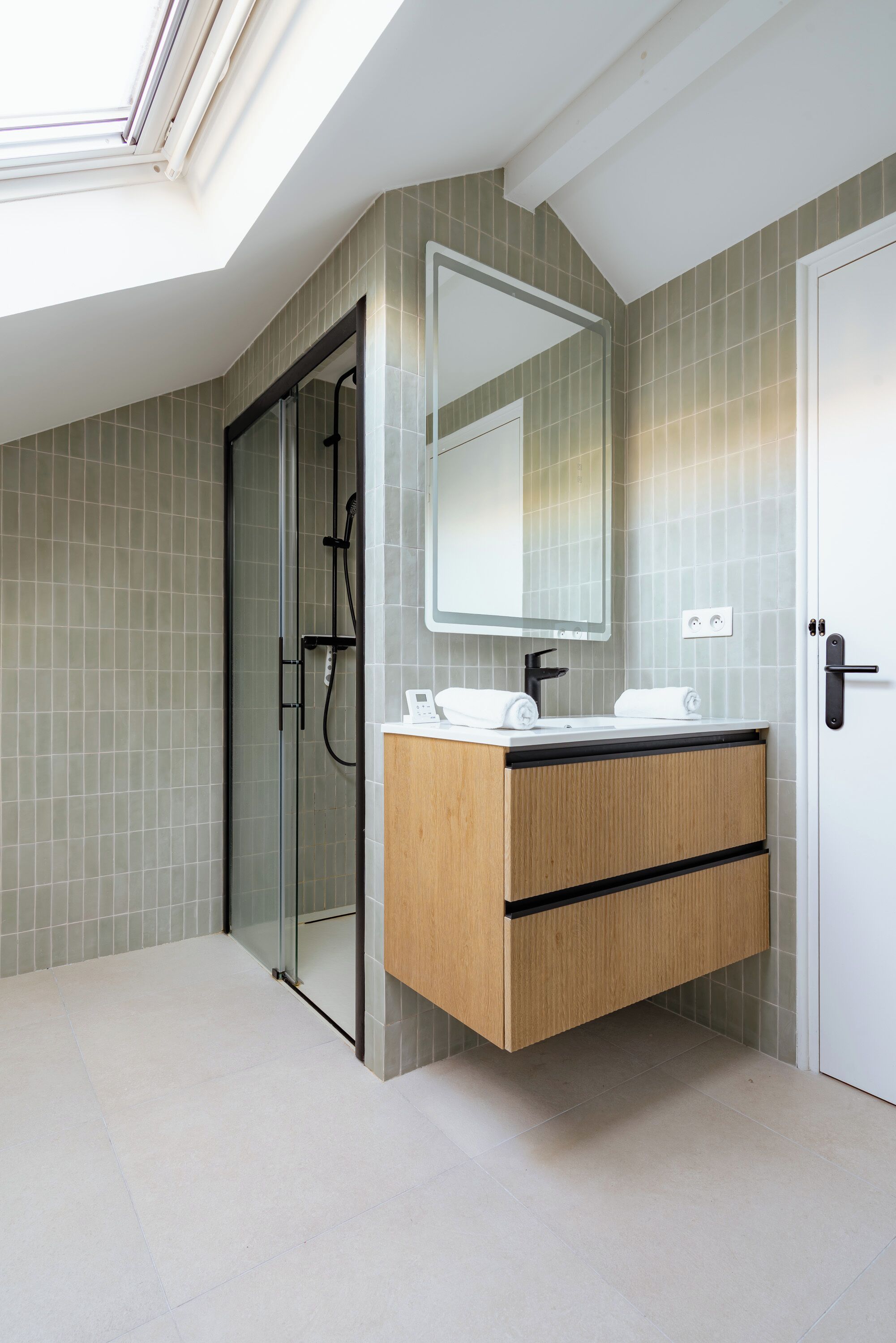 Une salle de bain moderne dotée d’un puits de lumière, d’une cabine de douche en verre, d’un meuble-lavabo en bois avec un miroir et d’une porte blanche.