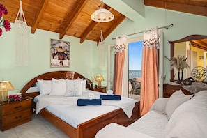Un dormitorio con techo de madera, una cama con ropa de cama blanca, una mesita de noche con lámpara y vistas al mar a través de la ventana.