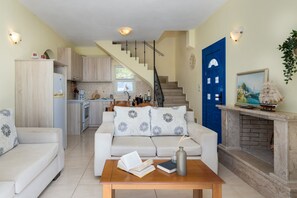 Uma sala de estar moderna com sofá, mesa de centro e lareira. Uma cozinha visível ao fundo apresenta armários de madeira e uma escada que leva a outro andar.