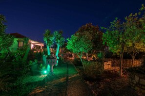 Un jardin nocturne avec des arbres illuminés et un petit bâtiment, entouré d’un mur de pierre et bordé d’un sentier.