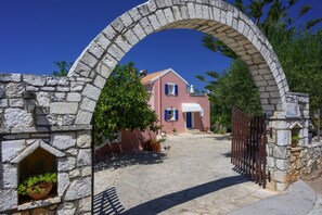 Uma arcada de pedra leva a um edifício cor de rosa com toldo branco, cercado por vegetação e um muro de pedra.