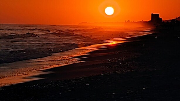 A sunset over a beach with waves, a distant city skyline, and a structure on the right. - Casa Ninì, charming accommodation in the center (Campofelice di Roccella)
