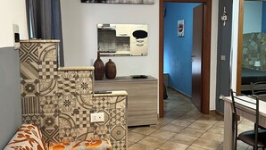 A living room with a patterned sofa, a dining area with a table and chairs, a kitchen area with a stove and cabinets, and a hallway leading to another room. - Casa Ninì, charming accommodation in the center (Campofelice di Roccella)