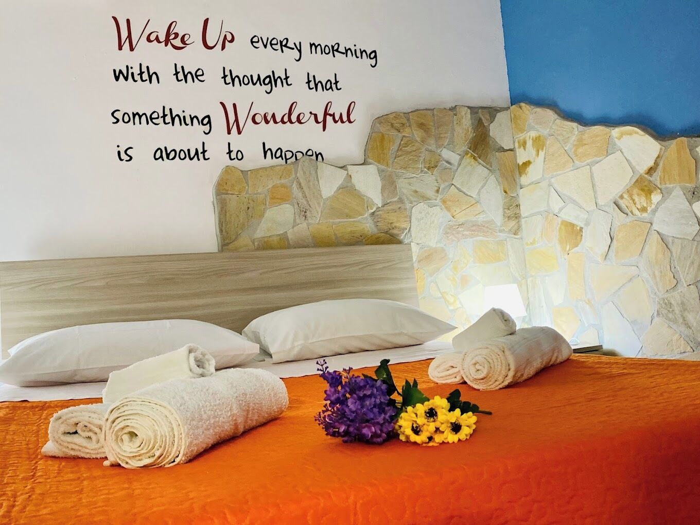 Ein ordentlich gemachtes Bett mit aufgerollten weißen Handtüchern, eine Steinsäule und ein motivierender Spruch an der Wand.