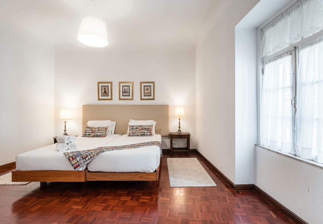 Un dormitorio con cama de madera, dos mesitas de noche, una ventana con cortinas translúcidas y obras de arte enmarcadas en la pared.