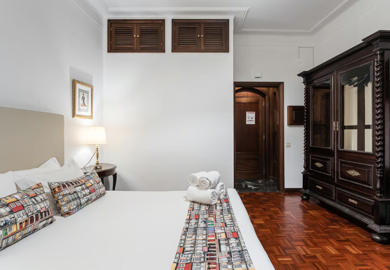 A hotel room with a large bed, a wooden wardrobe, a framed picture on the wall, and a door leading to another room.