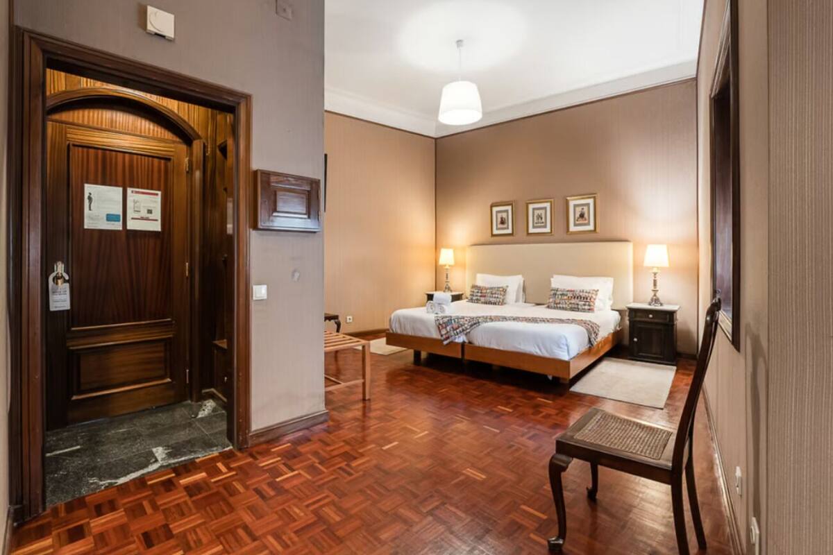 Een hotelkamer met een houten vloer, een tweepersoonsbed, twee nachtkastjes, een stoel en een deur met een bordje.